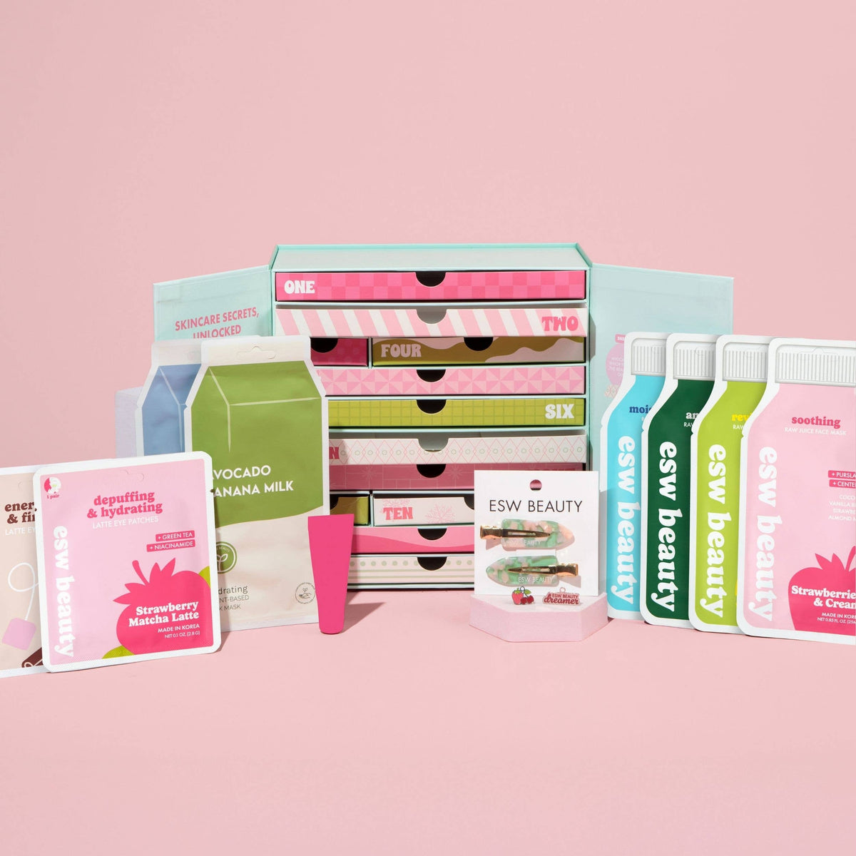 A skincare set display featuring multiple ESW Beauty products including a stack of drawer-style boxes, individual sachets, and a small tube, arranged on a solid pink background.