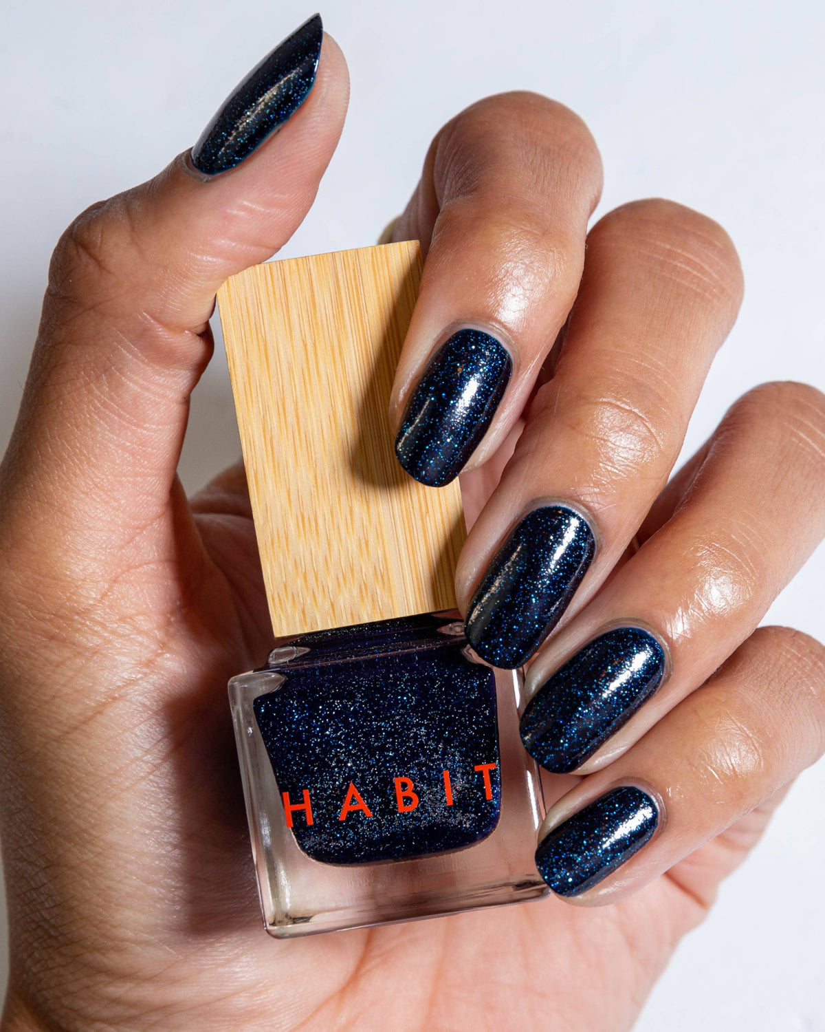 Hand with glittery blue nail polish holding a bottle of 'Habit' nail polish.