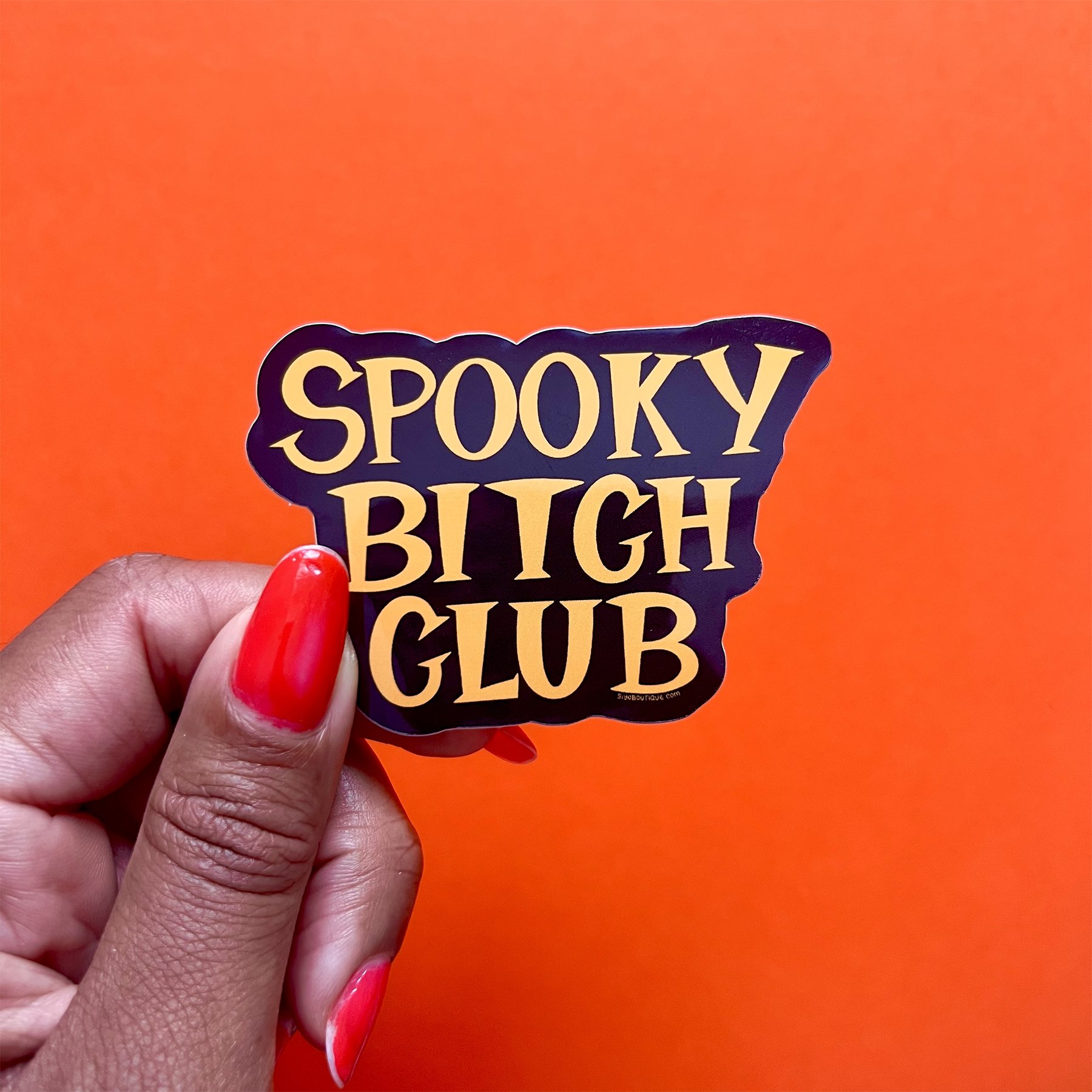 Hand holding a sticker with 'Spooky Bitch Club' text against an orange background