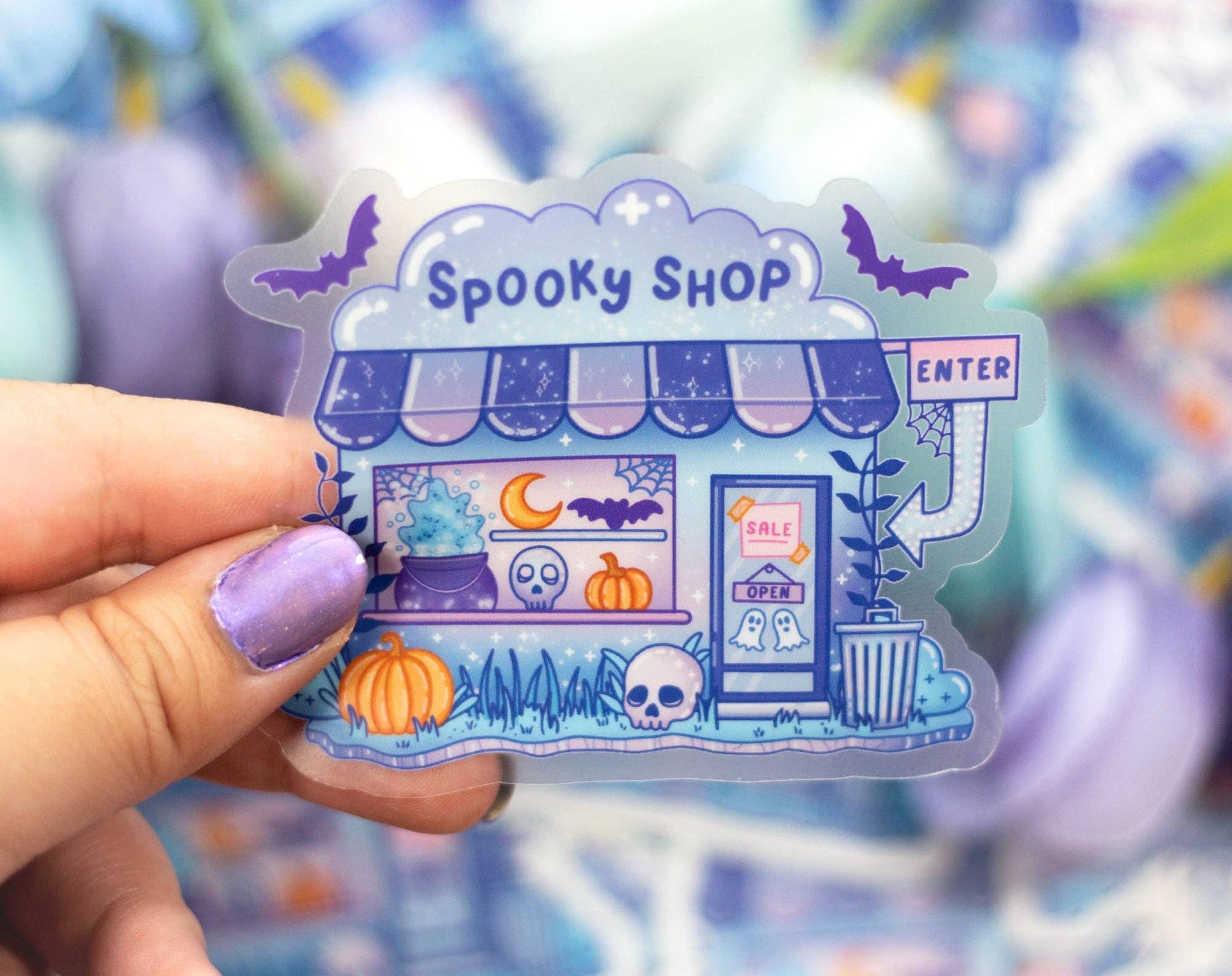 Halloween-themed sticker of a 'Spooky Shop' held by a hand with purple nail polish.