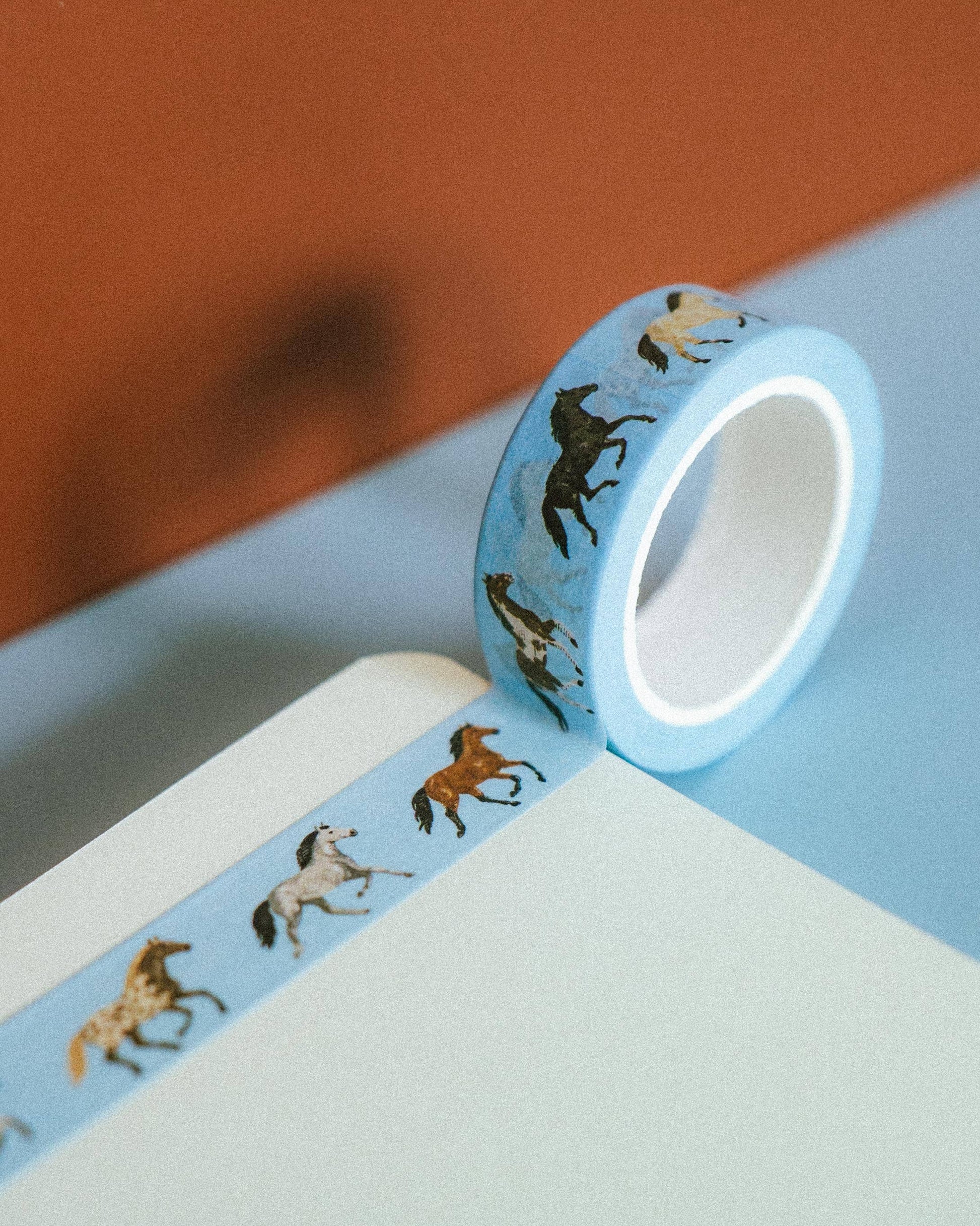 A roll of light blue washi tape with a pattern of running horses in black, brown, and white, shown partially unrolled on a light blue surface against a warm terracotta background.