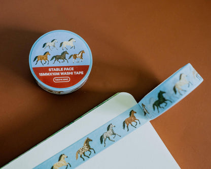 A roll of washi tape with a light blue background and a pattern of running horses in black, brown, and white, shown next to an open notebook on a warm brown surface.