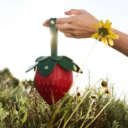 Strawberry-shaped bag held by a hand in a natural setting with flowers.