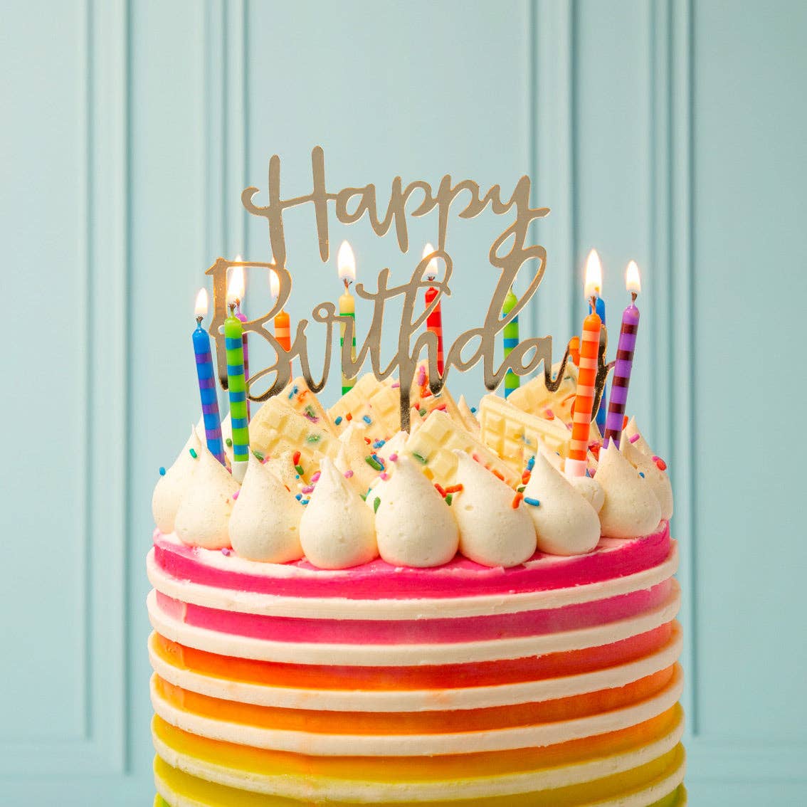 Colorful birthday cake with lit candles and 'Happy Birthday' topper against a light blue background