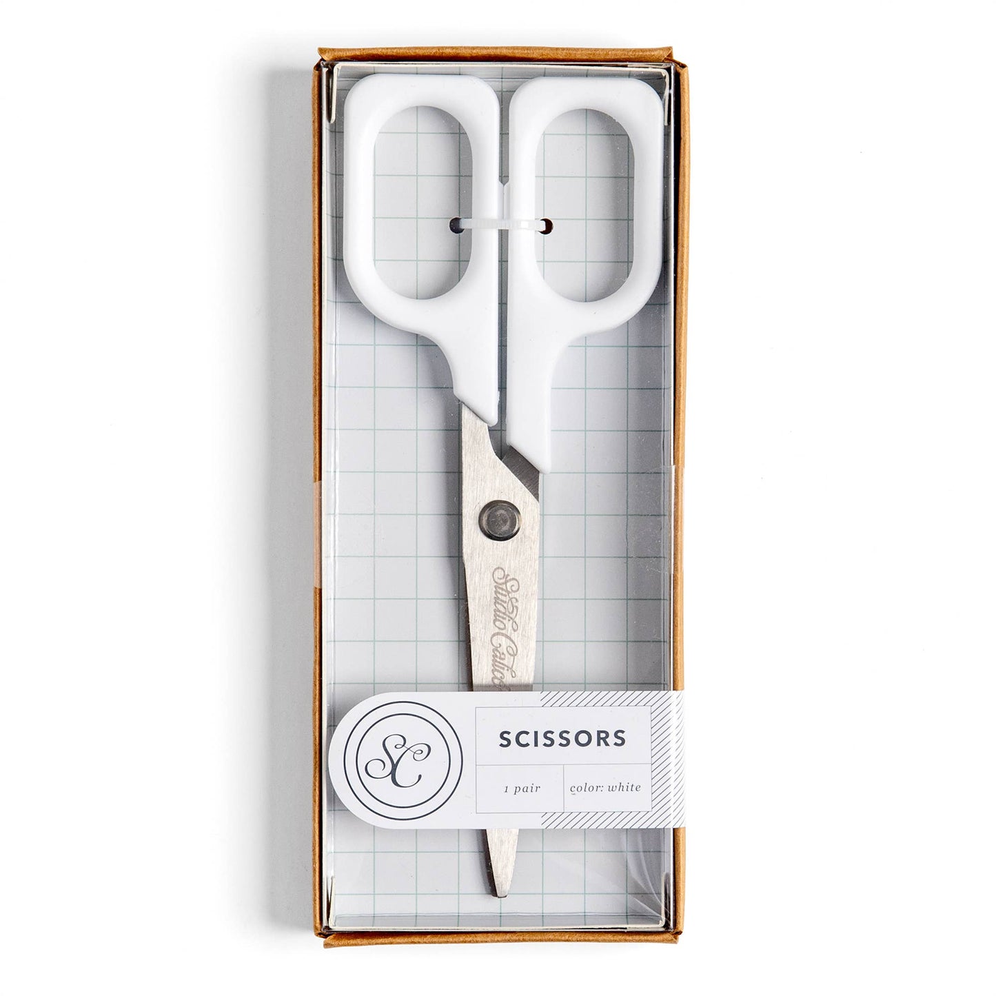 White-handled scissors in a clear-front gift box with a white paper label that reads 'SCISSORS' and shows a monogram logo.