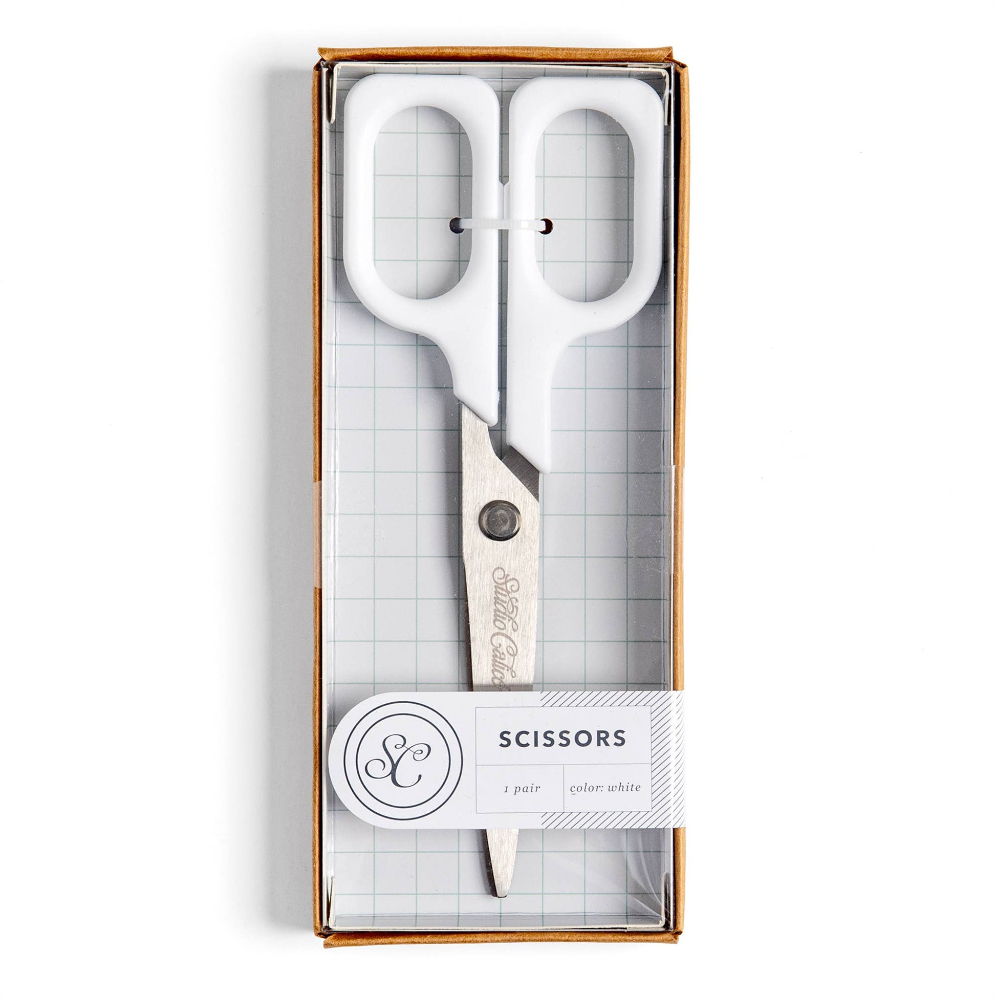 White-handled scissors in a clear-front gift box with a white paper label that reads 'SCISSORS' and shows a monogram logo.