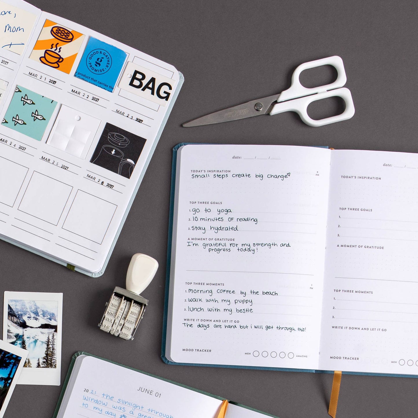 An open notebook with handwritten notes and trackers lies on a dark gray surface alongside a closed printed planner or calendar, a pair of white-handled scissors, a small white eraser, metal paper clips, and a few small printed cards.