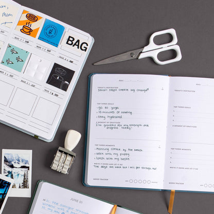 An open notebook with handwritten notes and trackers lies on a dark gray surface alongside a closed printed planner or calendar, a pair of white-handled scissors, a small white eraser, metal paper clips, and a few small printed cards.