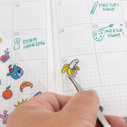 Calendar with colorful stickers and a hand using tweezers to place a banana sticker.