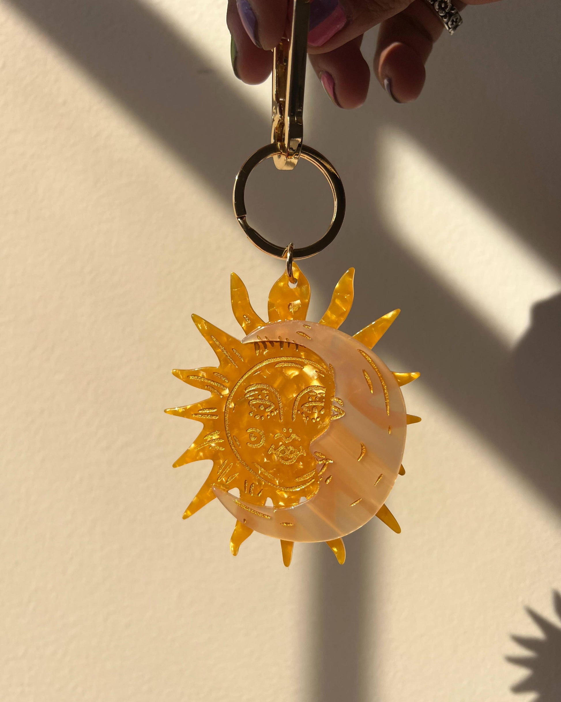 A translucent amber-colored acrylic keychain featuring a stylized sun and crescent moon design, held by a hand against a warm beige wall with strong sunlight and shadows.