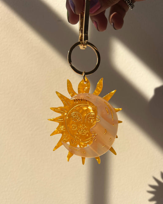 A translucent amber-colored acrylic keychain featuring a stylized sun and crescent moon design, held by a hand against a warm beige wall with strong sunlight and shadows.
