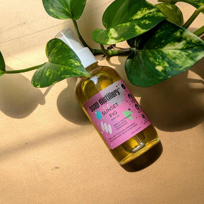 Bottle of 'Sunset Fig' from Soap Distillery on a beige surface with green leaves.