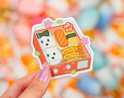 Sticker of a bento box with a dog and food items held by a hand against a colorful blurred background. Sushi Sticker