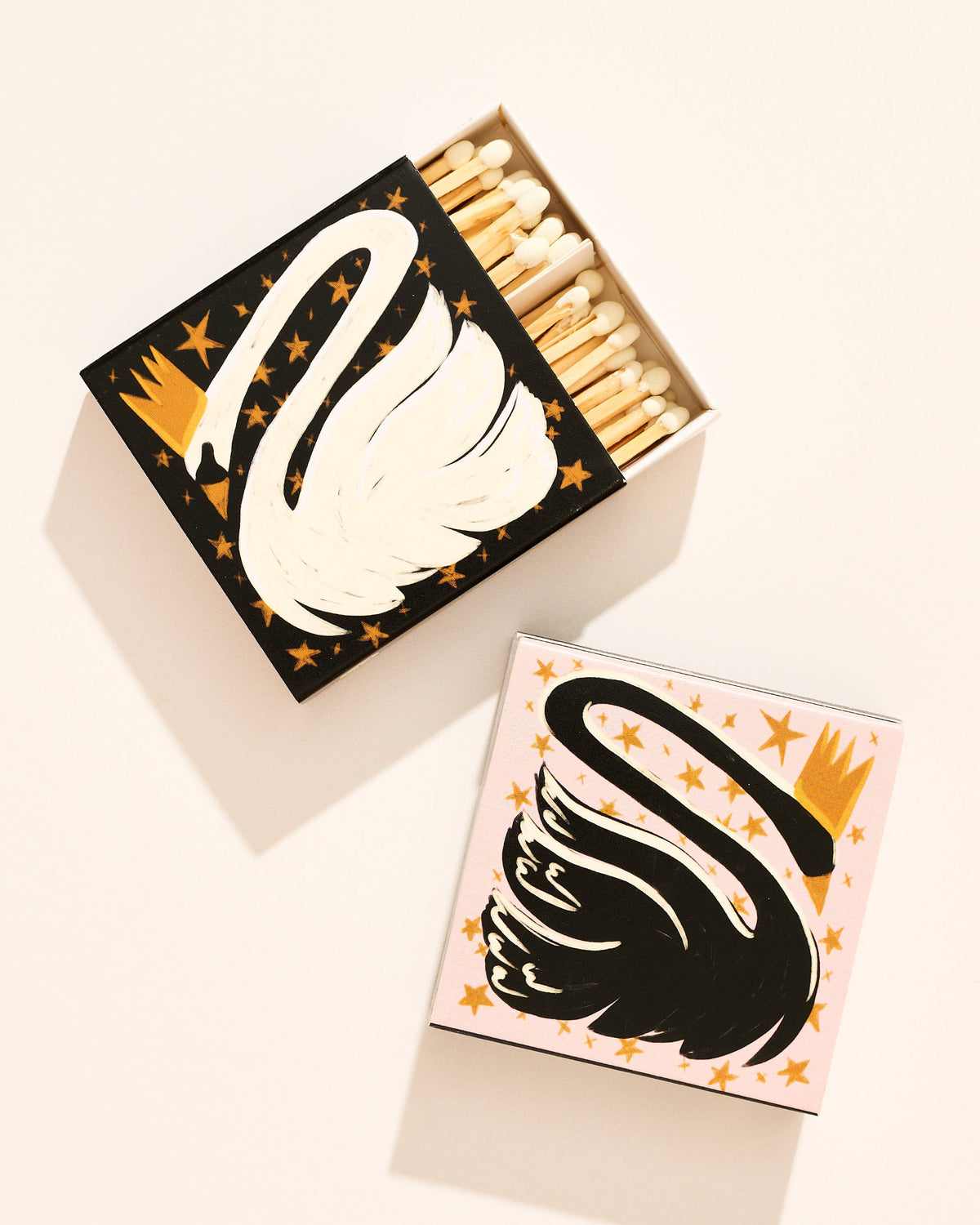 Two decorative matchboxes with a stylized swan and gold stars, one black-and-white and one pink-and-black, shown open with matches inside on a warm off-white background.