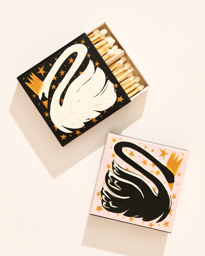 Two decorative matchboxes with a stylized swan and gold stars, one black-and-white and one pink-and-black, shown open with matches inside on a warm off-white background.