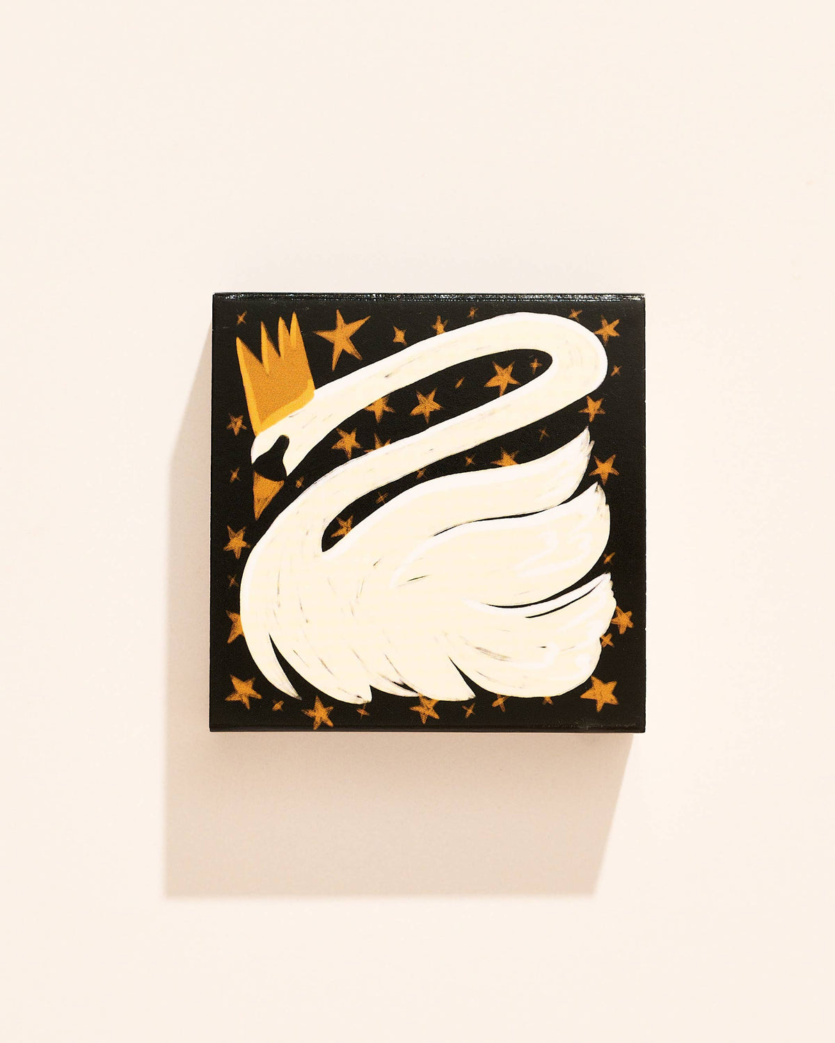 A square matchbox featuring a stylized white swan with a gold crown, surrounded by small gold stars, photographed against a warm off-white background.