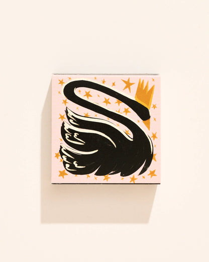 A square matchboxfeaturing a stylized black swan with a gold crown on a pale pink background with scattered gold stars, photographed against a warm off-white wall.