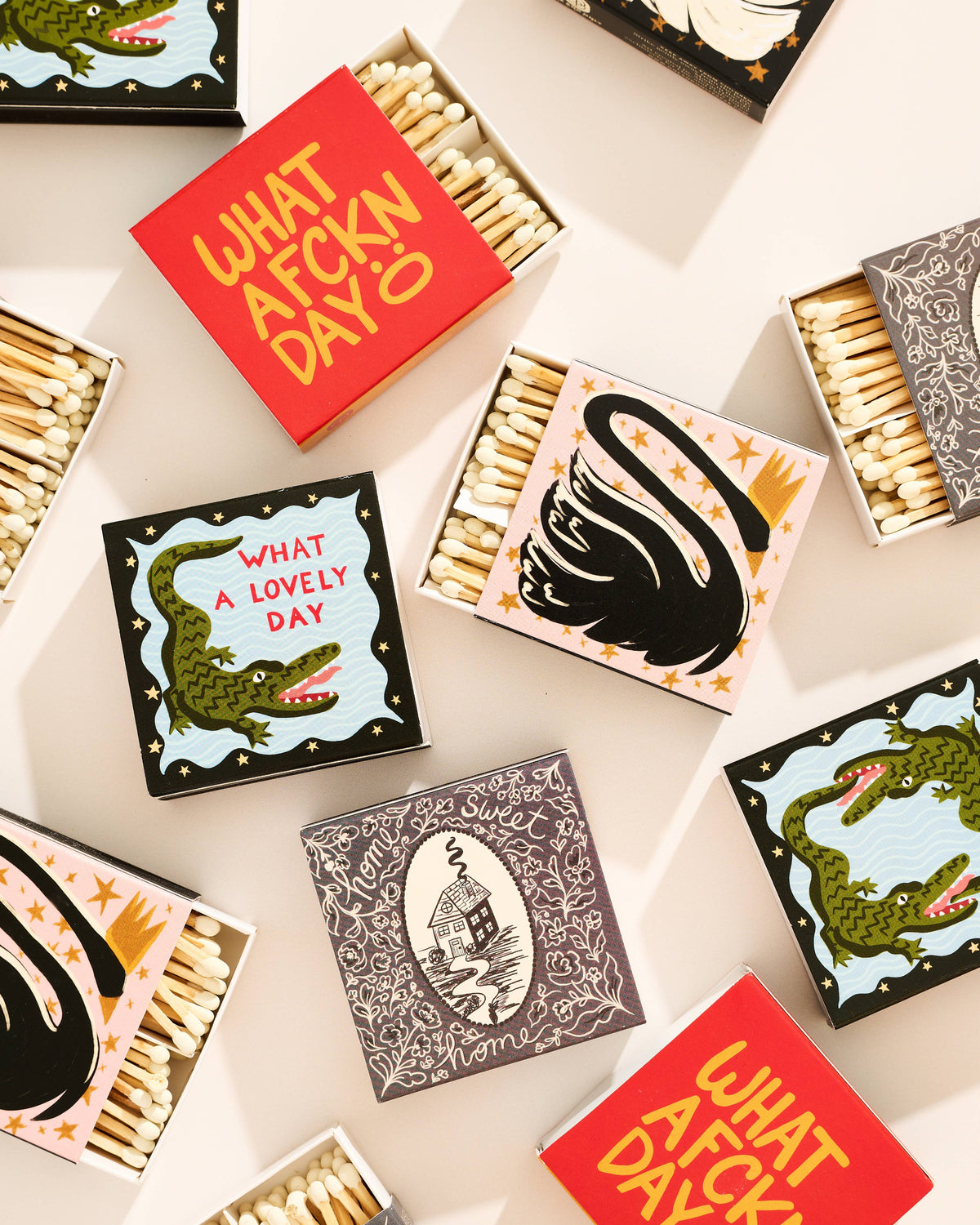 A styled flat-lay of decorative matchboxes arranged on a light beige background. Several open matchboxes reveal beige wooden matches, while closed boxes feature colorful illustrated lids with phrases like “WHAT A F*CK N DAY” and “WHAT A LOVELY DAY,” along with black swan and green crocodile illustrations.
