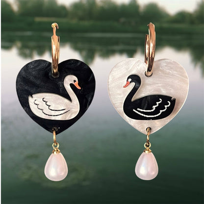 Heart-shaped earrings with swan designs and pearl drops against a blurred natural background