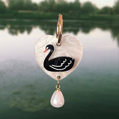 Heart-shaped pendant with swan design and pearl drop against a blurred natural background