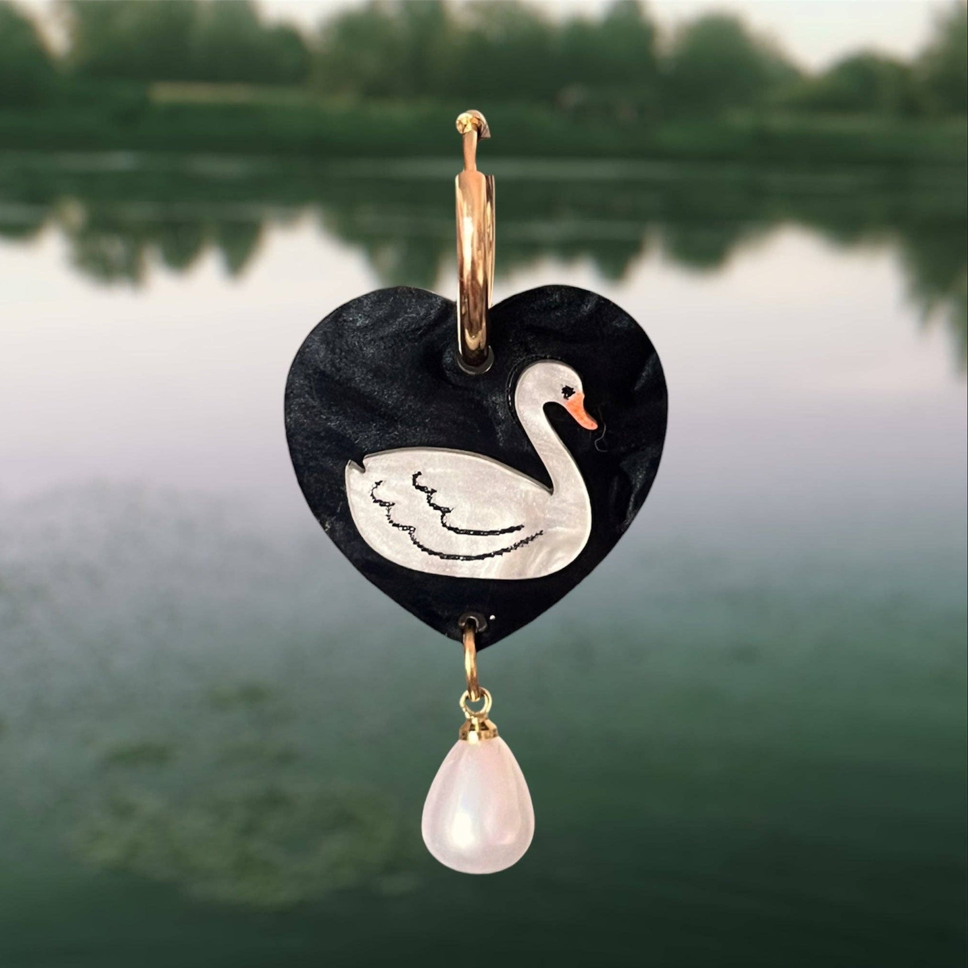 Heart-shaped pendant with a swan design and pink teardrop charm against a blurred natural background