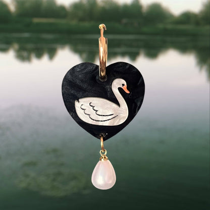 Heart-shaped pendant with a swan design and pink teardrop charm against a blurred natural background