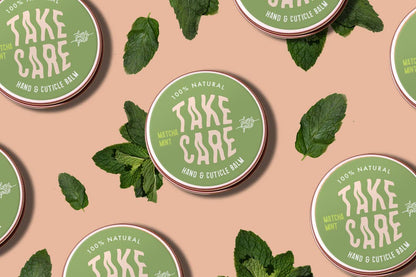 Green containers of 'Take Care' hand and cuticle balm with mint leaves on a beige background