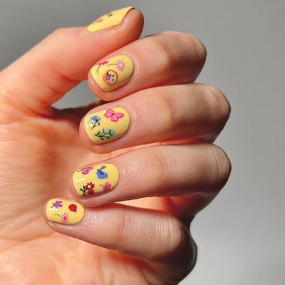 Hand with yellow nail polish featuring floral and butterfly designs on a neutral background