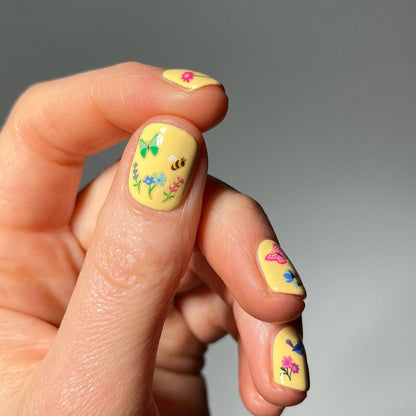 Hand with yellow nail polish featuring floral and butterfly designs on a gray background