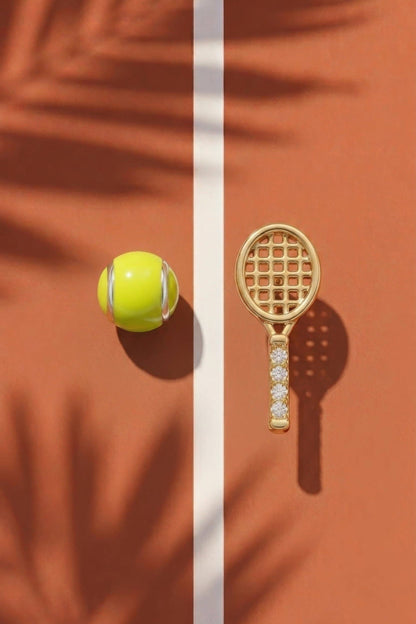 A gold tennis racket and tennis ball set displayed on a terracotta court with a white center line and palm leaf shadows.