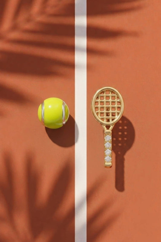A gold tennis racket and tennis ball set displayed on a terracotta court with a white center line and palm leaf shadows.