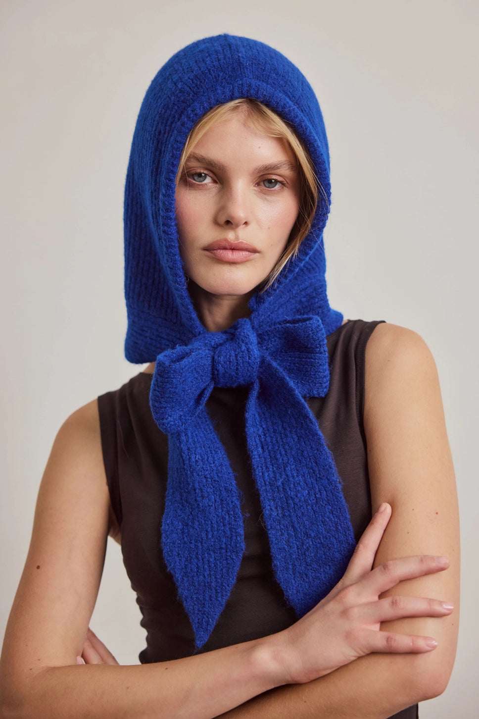 A woman wearing a bright royal blue knitted bonnet with a large bow, styled over a dark brown sleeveless top against a plain light beige studio background.