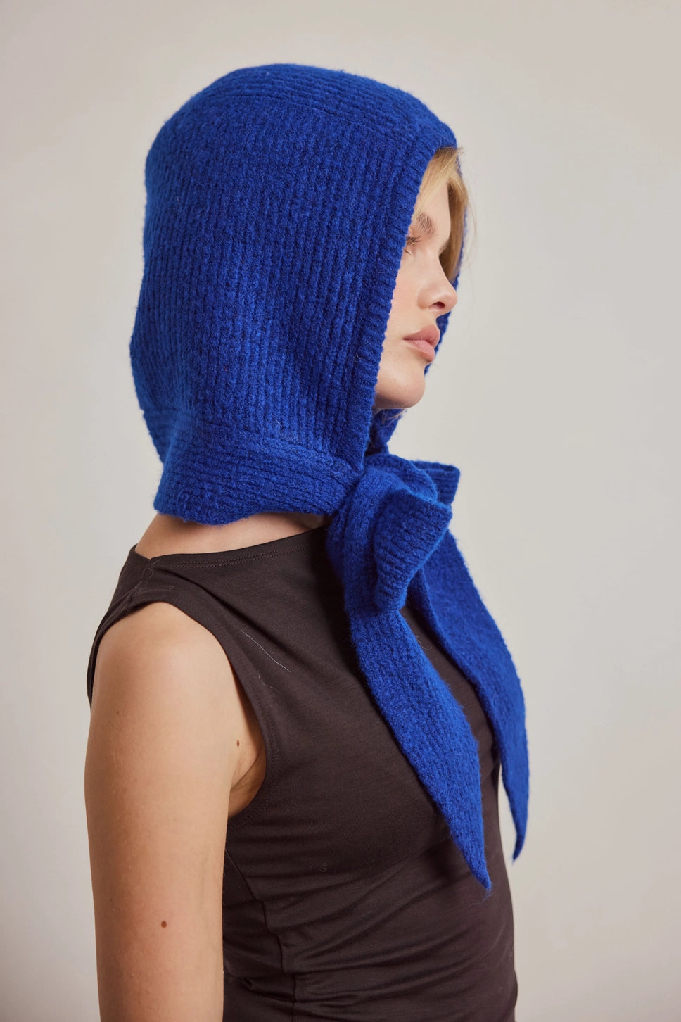 A woman shown in side profile wearing a bright royal blue knitted bonnet with a long tail that drapes down the back, paired with a dark brown sleeveless top against a plain light gray background.