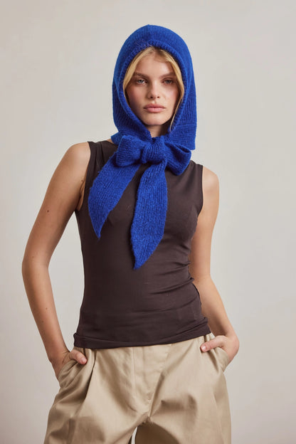 A model wearing a vibrant royal blue knitted bonnet with a large bow, styled over a dark brown sleeveless top and beige trousers, set against a plain light gray studio background.