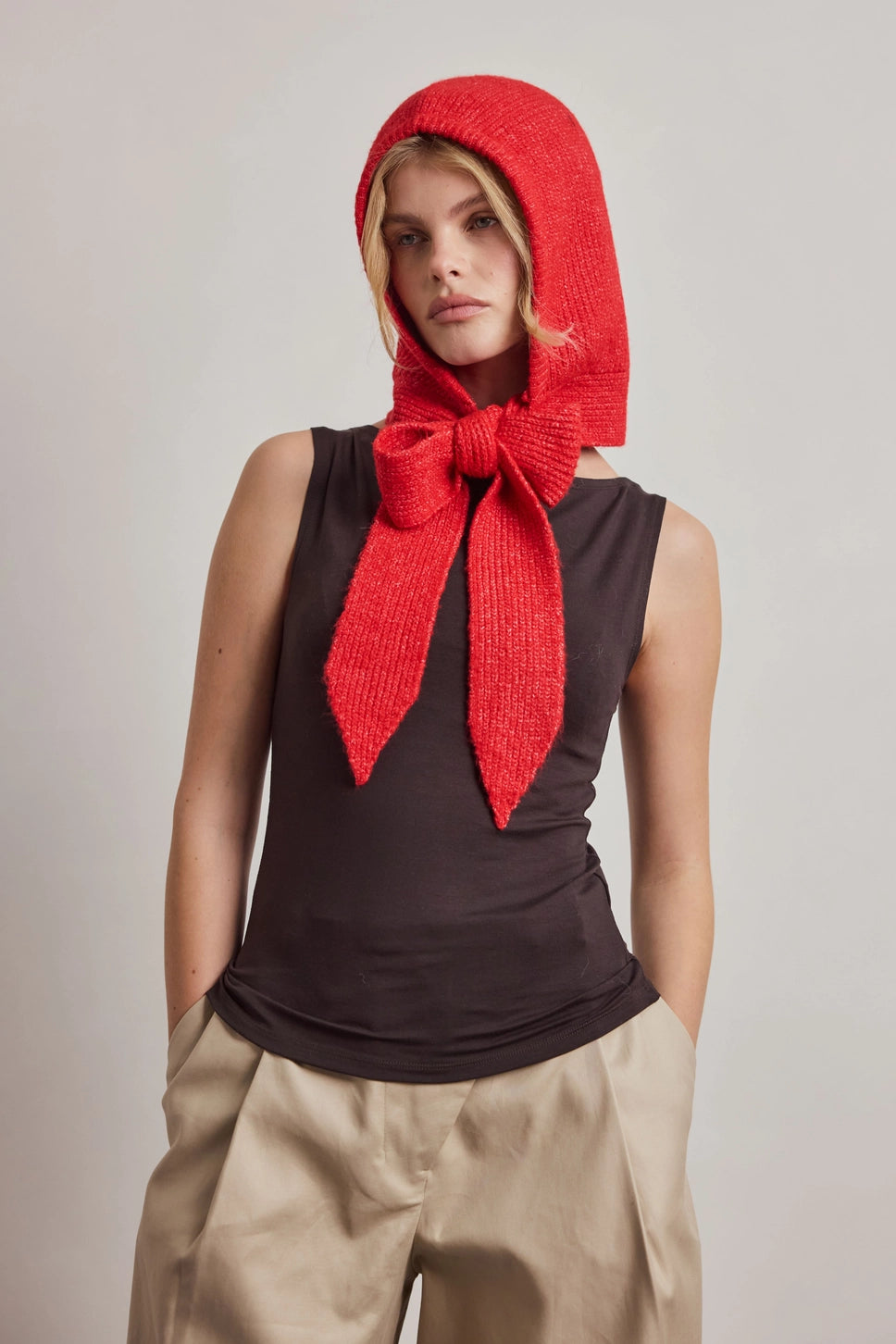 A model wearing a bright red textured knit bonnet with a large bow, styled over a dark brown sleeveless top and beige trousers, set against a plain light gray studio background.
