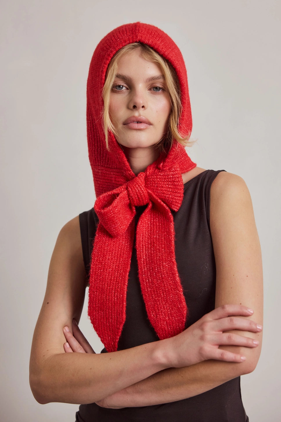 A model wearing a vibrant red knit bonnet with a long tail that ties into a large bow, styled over a dark brown sleeveless top against a neutral light gray background.