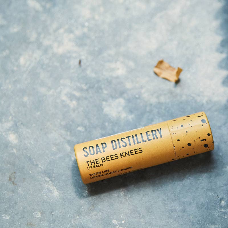 Lip balm container labeled 'The Bees Knees' from Soap Distillery on a textured surface.