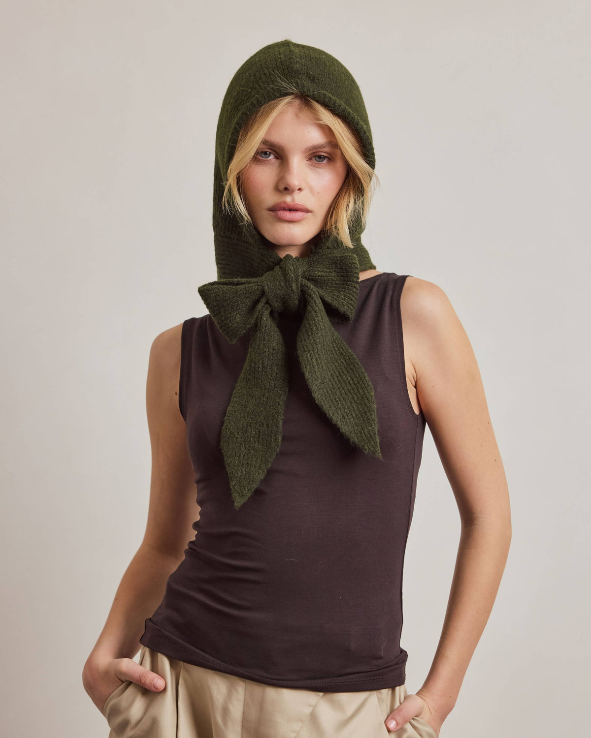 A model wearing an olive green knit hood with a large bow at the neck, styled with a dark brown sleeveless top and beige trousers against a neutral studio background.