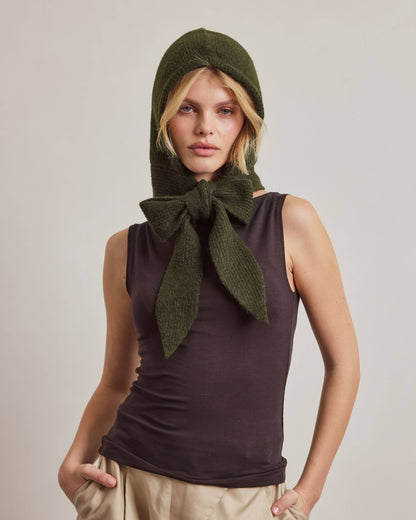 A model wearing an olive green knit hood with a large bow at the neck, styled with a dark brown sleeveless top and beige trousers against a neutral studio background.
