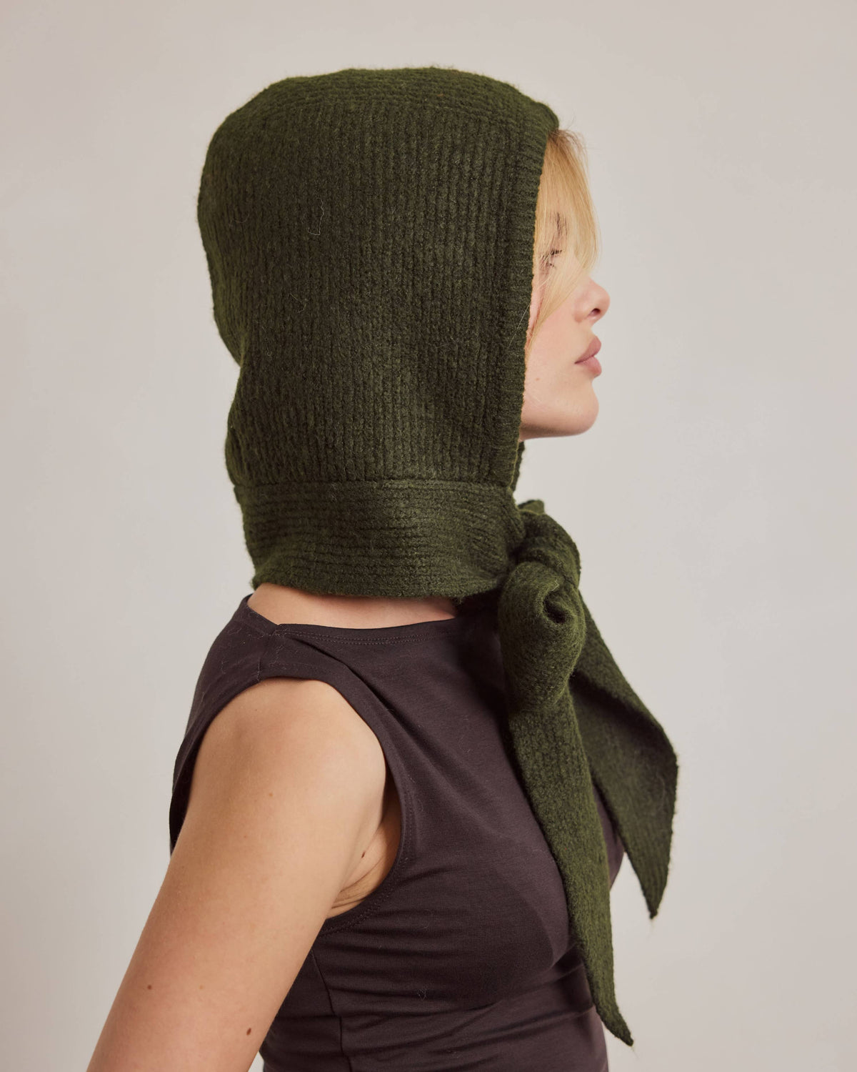 A woman in profile wearing a dark olive green ribbed knit bonnet with a large bow, paired with a dark brown sleeveless top, against a plain light gray background.