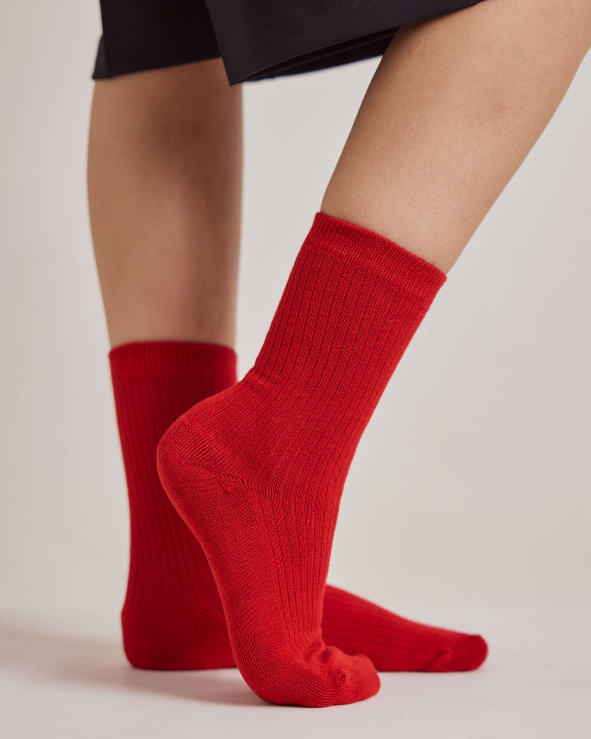 A close-up of a person's feet wearing bright red ribbed crew socks, with the lower legs of a black skirt visible against a light gray studio background.