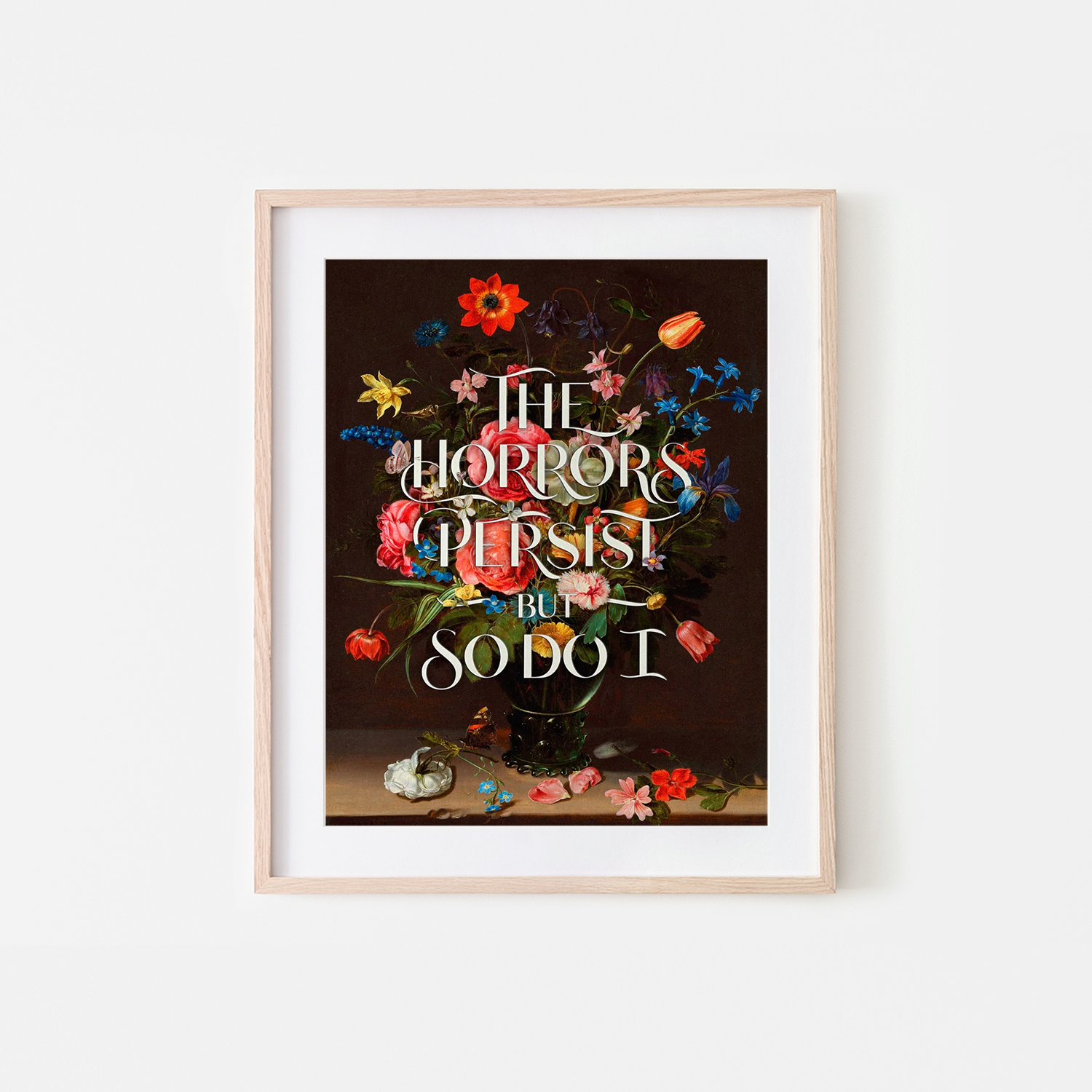 A framed art print featuring a dark floral still life with the text 'THE HORRORS PERSIST BUT SO DO I' in ornate white lettering, displayed against a light gray wall.