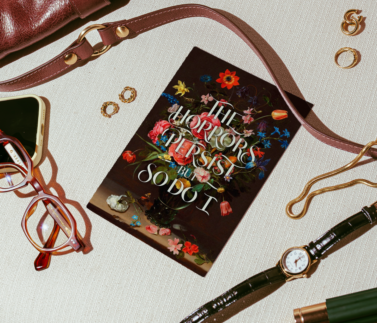 A styled flat-lay featuring a book titled 'The Horrors Persist So Do I' with a dark floral cover, surrounded by fashion accessories including a brown leather shoulder bag, eyeglasses, rings, a watch, and a pen on a textured cream fabric background.