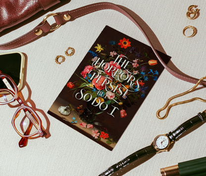 A styled flat-lay featuring a book titled 'The Horrors Persist So Do I' with a dark floral cover, surrounded by fashion accessories including a brown leather shoulder bag, eyeglasses, rings, a watch, and a pen on a textured cream fabric background.