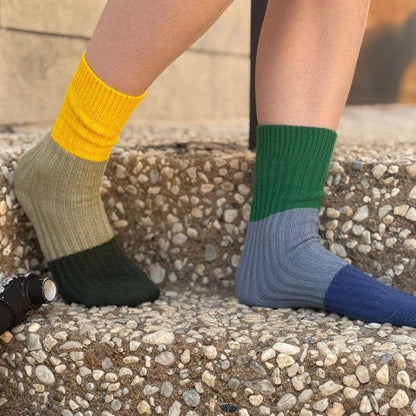A person wearing three-color ribbed crew socks on their feet, standing on outdoor concrete steps. One sock is yellow and olive green, the other is green and blue-gray, and the third is blue.