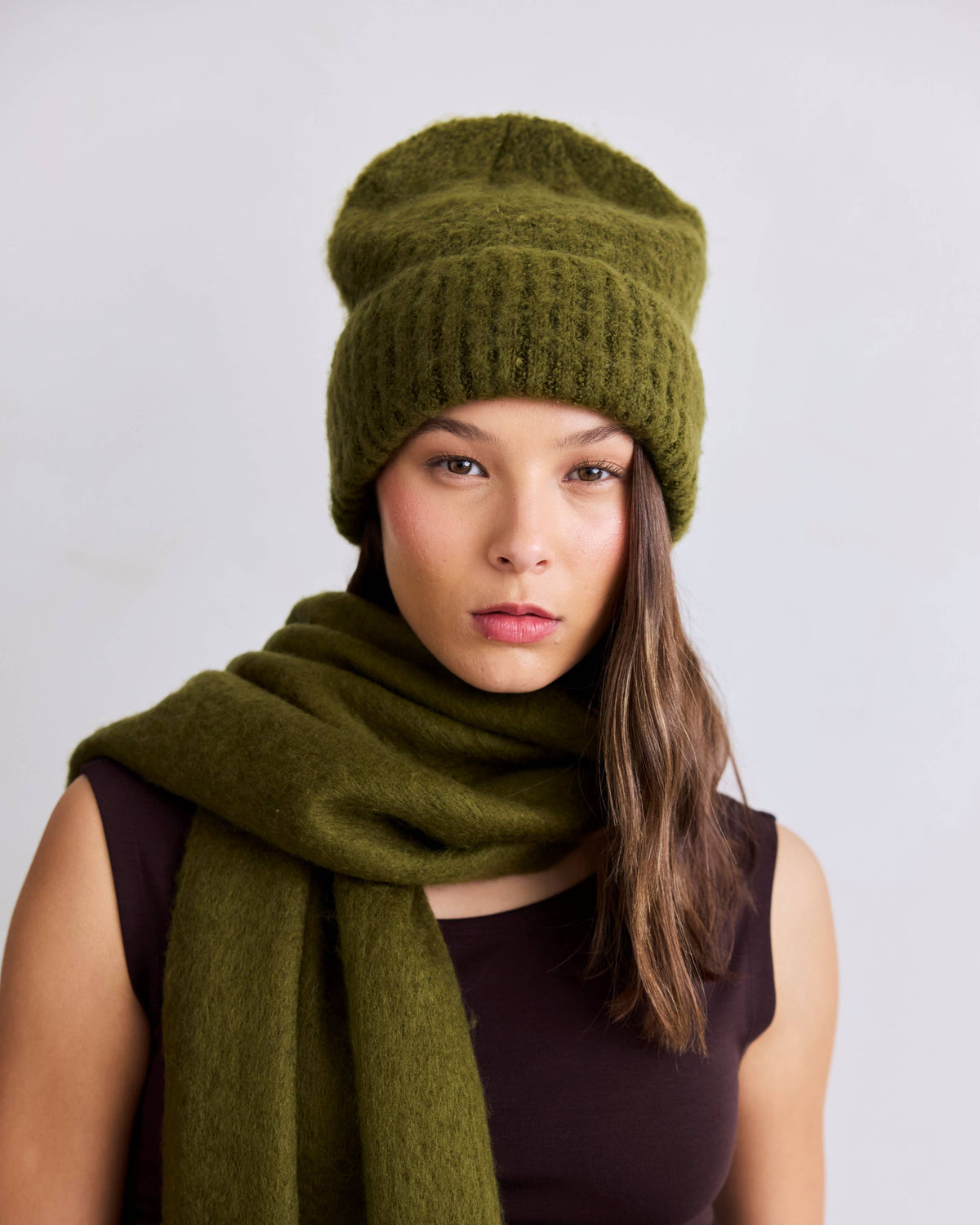 A woman wearing an olive green knit beanie and a matching olive green scarf, paired with a dark sleeveless top, against a plain light gray studio background.