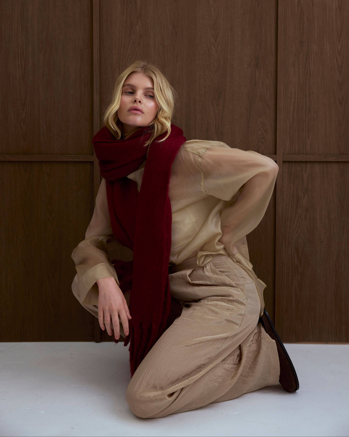 A woman kneels on a light floor against a dark wood-paneled wall, wearing a beige sheer long-sleeve top and matching beige pants with a deep burgundy scarf.