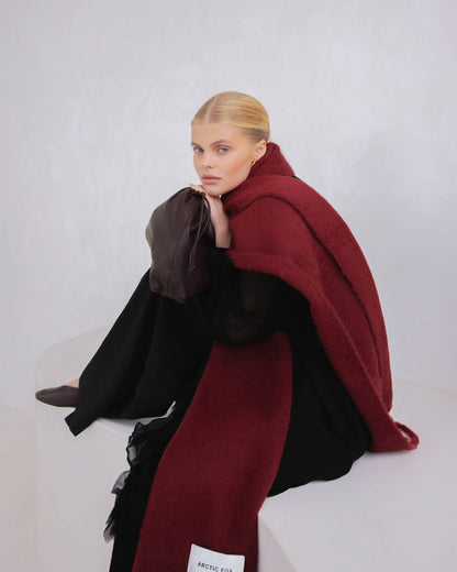 A blonde model sits on a white platform wearing a long dark brown wrap and a large deep burgundy scarf draped over her shoulders. A small white label at the bottom of the scarf reads Arctic Fox