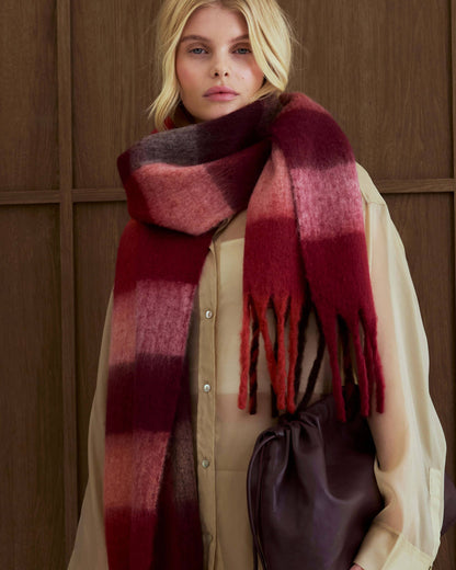 A woman wearing a thick, oversized gradient scarf in shades of burgundy, red, and dark brown, styled over a cream-colored button-up shirt and holding a dark brown leather drawstring bag against a dark wood-paneled background.