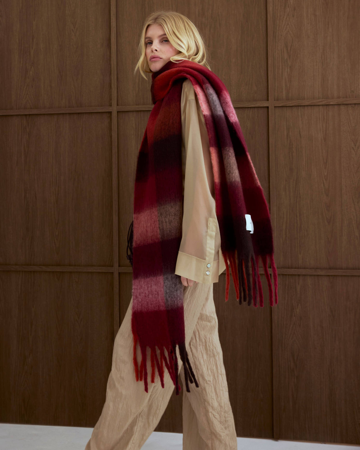 A blonde model wearing a thick, striped plaid scarf in shades of red, burgundy, and black, styled over a beige top and beige trousers, standing against a dark wood-paneled wall.